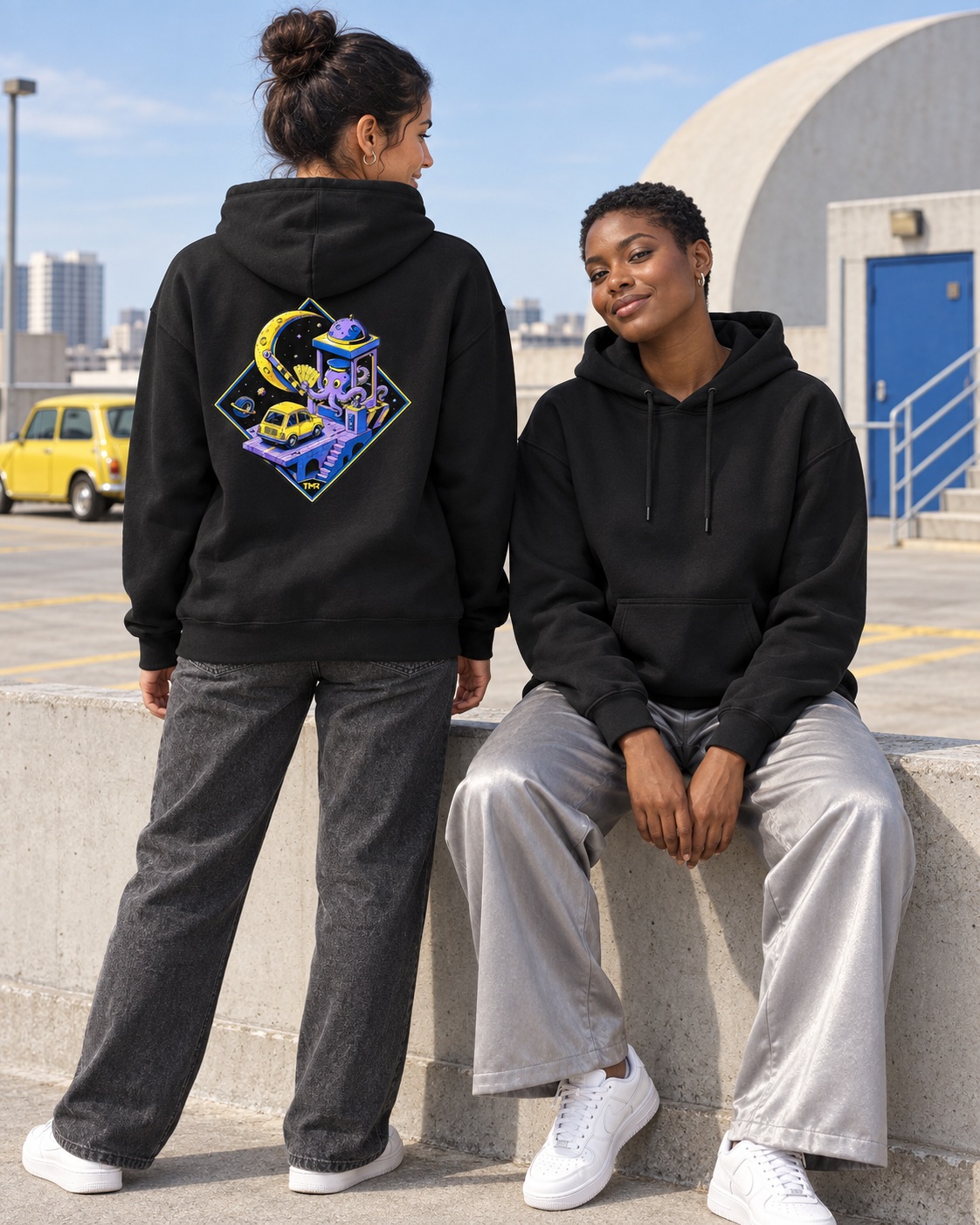 Peaje lunar — Sudadera 4 Dos modelos adultas ficticias con la sudadera negra Peaje lunar de TMR en un aparcamiento urbano de día: frontal liso y espalda con pulpo, coche amarillo y peaje espacial en violeta y azul.