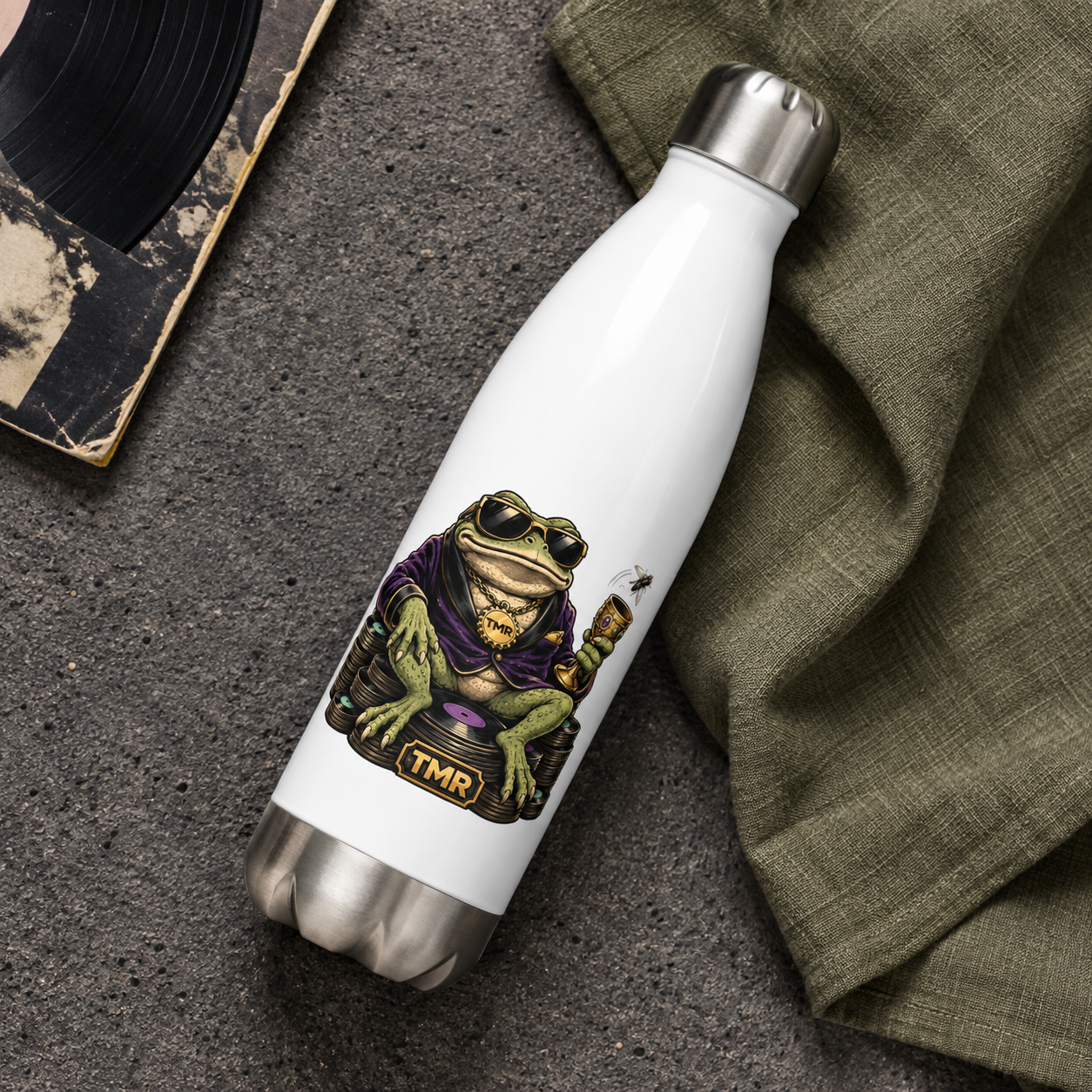Sapo millonario — Botella térmica 500 ml 3 Vista cenital de la botella térmica Sapo millonario de TMR sobre piedra oscura y tela verde, con el estampado frontal visible.
