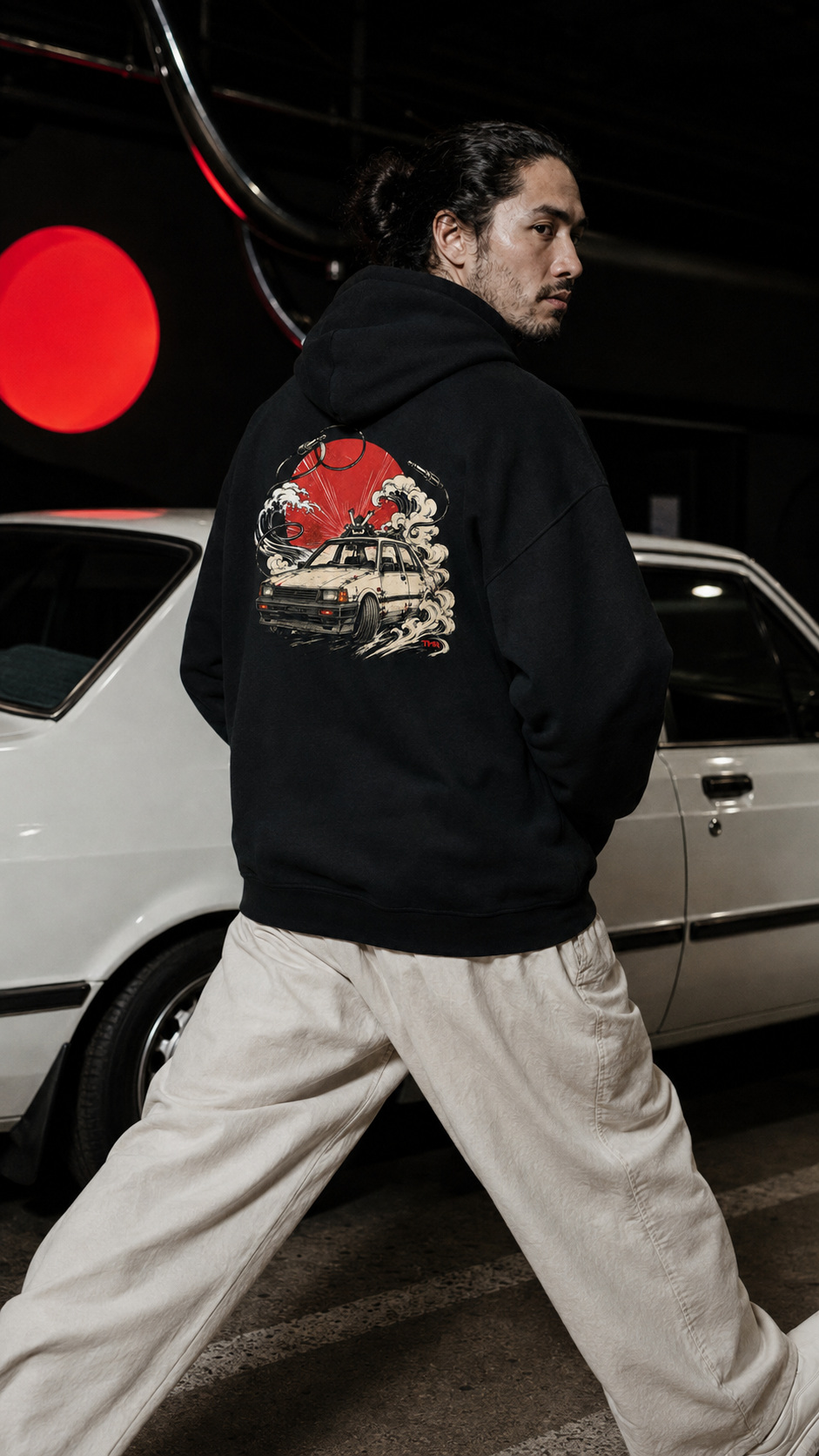 Sedán samurái — Sudadera 3 Mockup digital de la sudadera negra Sedán samurái de TMR by Tamar Melián, vista de espalda con coche clásico, olas y círculo rojo, junto a un vehículo.