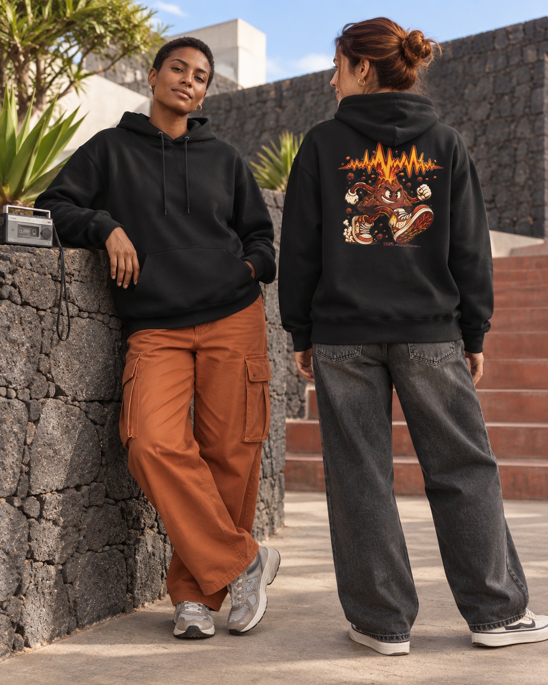 Volcán en zapatillas — Sudadera 4 Dos modelos adultas ficticias con la sudadera negra Volcán en zapatillas de TMR en un patio de piedra volcánica de día: frontal liso y espalda con volcán ilustrado corriendo con zapatillas.